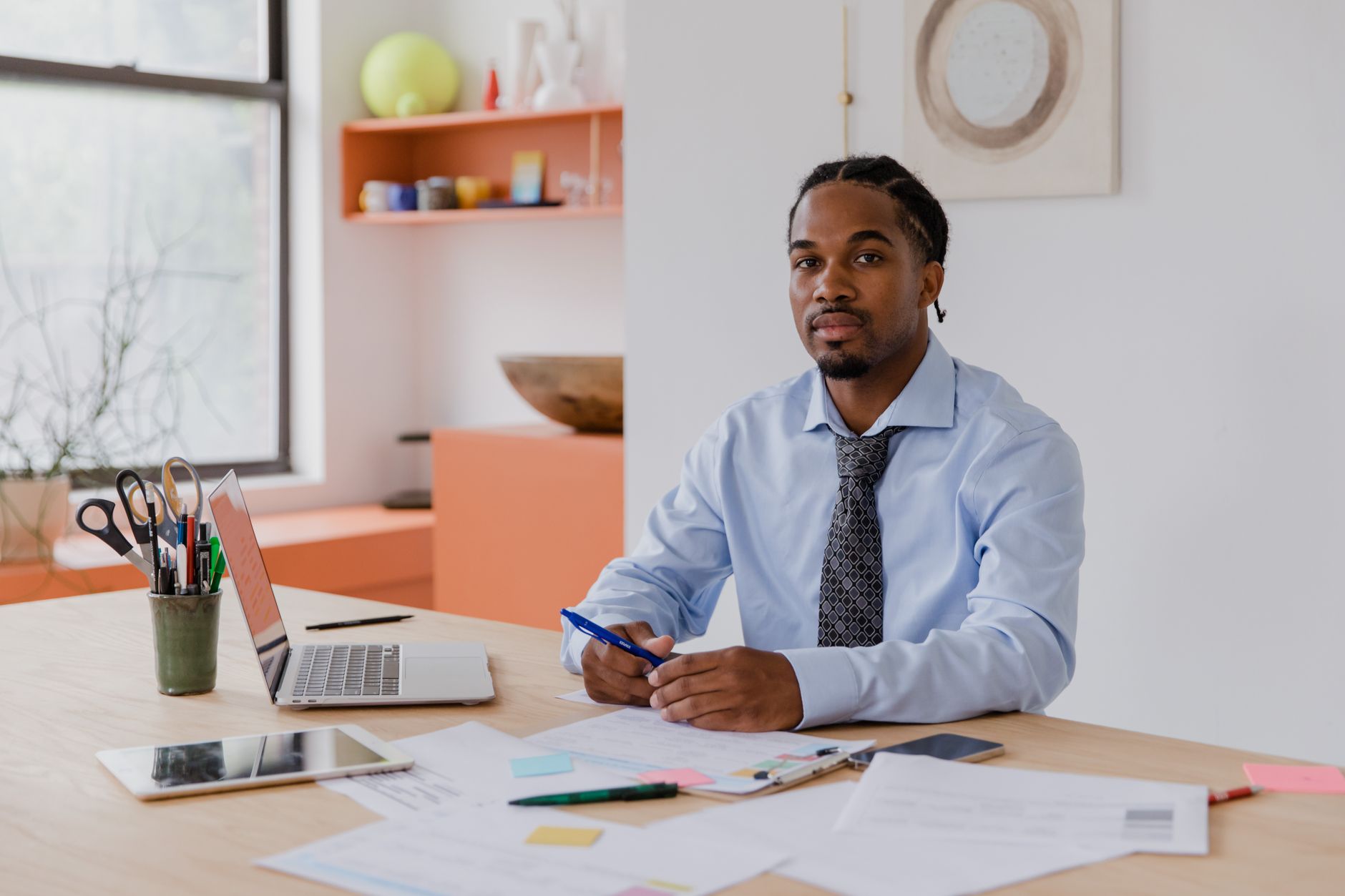 A professional businessman with a beard and braided hair sits at his desk with a laptop and documents, working diligently. —