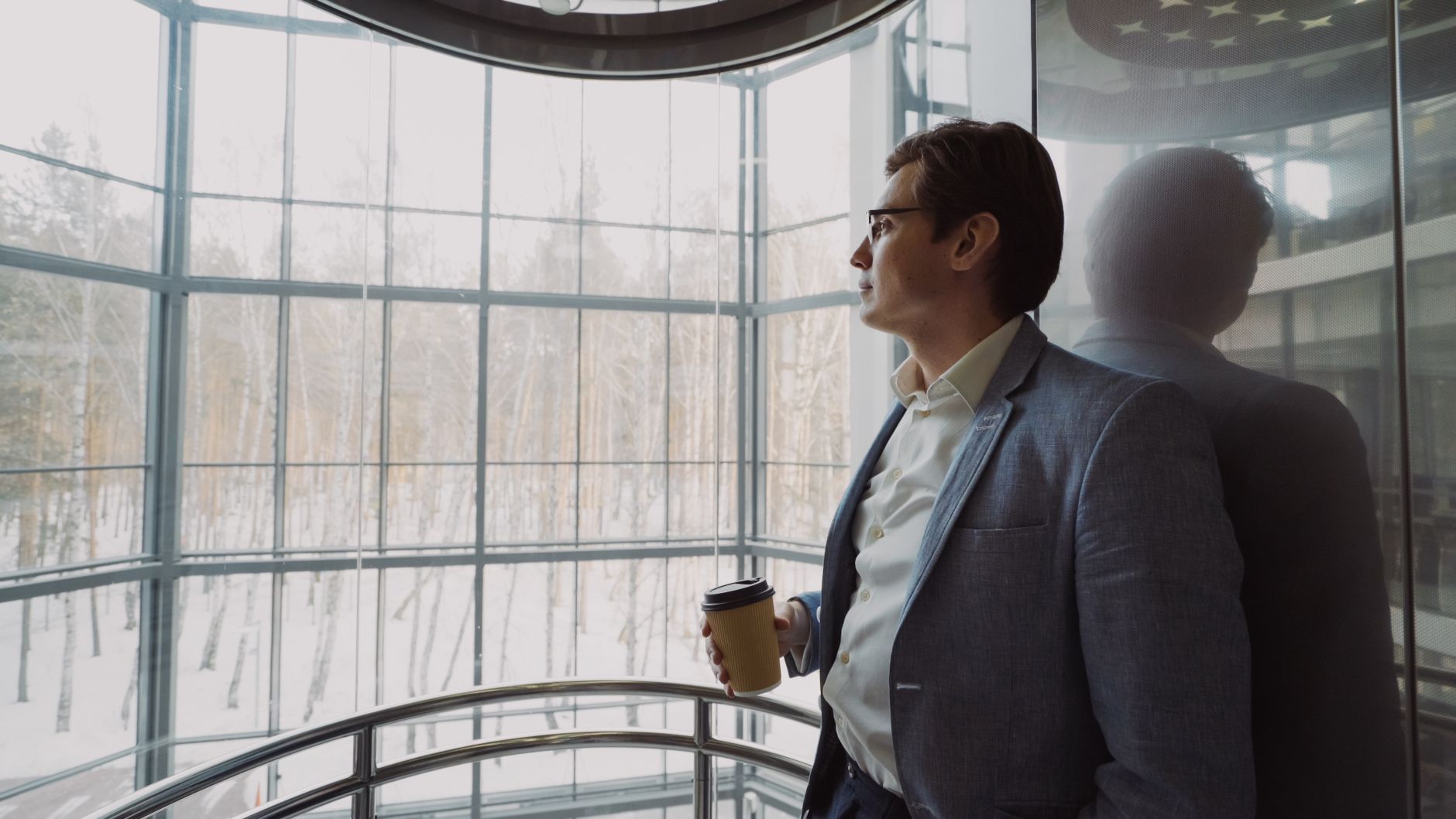 Businessman in blue suit with coffee cup looking out a large window, reflecting on a winter landscape. — Forget About Excuses