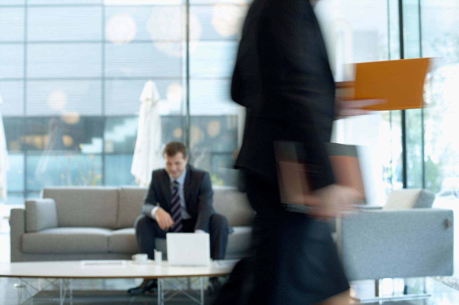 Businessman sitting in modern office lounge using a laptop while colleague walks past, creating dynamic blur. — Full details
