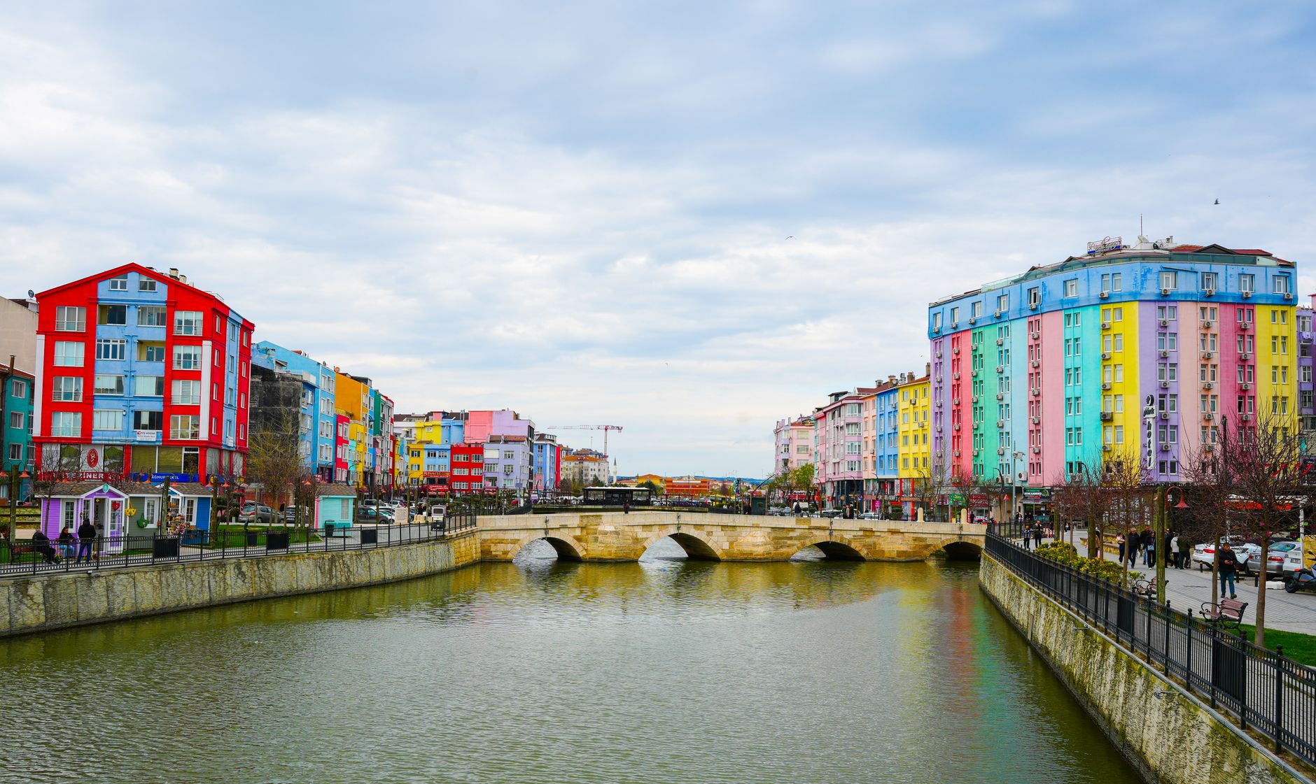 Vibrant cityscape with colorful buildings along a river and stone bridge under a cloudy sky. — Optimized Writing: Using The C