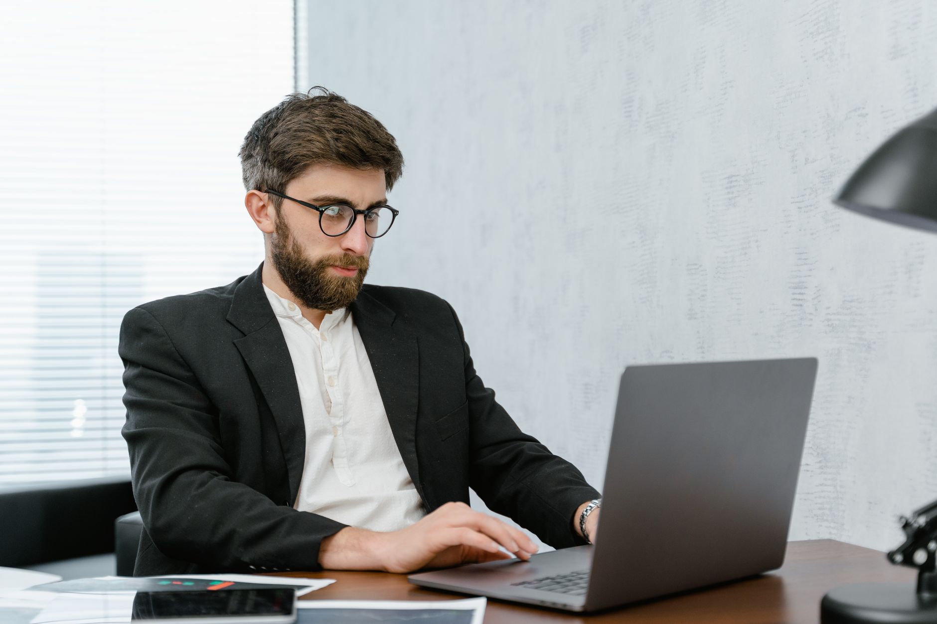 A bearded man in glasses and suit working on a laptop at an office desk. — Responsibility of Entrepreneurs