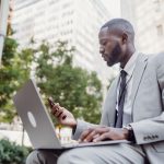 Businessman in a suit using a laptop and smartphone in an urban setting, blending technology with business. — Top Forex Tradi